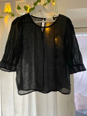 Svart transparent blus - Svart blus i transparent, lätt material med rund halsringning och volang trekvartsärmar. Blusen har en enkel design med liten knapp i nacken och volangdetaljer vid ärmslut. Perfekt för att skapa en cool och trendig look.