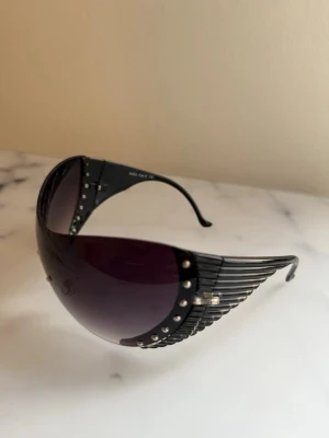 Y2K 2000s solglasögon med nitar - Säljer ett par Y2k svarta solglasögon med futuristisk sköldform och silvriga nitar längs sidorna. Glasen är mörka och tonade, bågarna har en unik ribbad design.