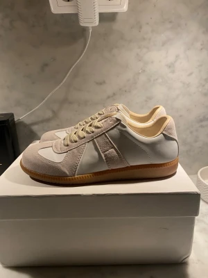 Vita och beige sneakers med mockadetaljer - Stilrena sneakers i vitt och beige med ovandel i läder och mocka, samt klassisk gummisula. Skorna har låga skaft och vita snören. Perfekta för dig som gillar minimalistisk och tidlös design.