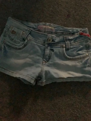 Ljusblå jeansshorts - Snygga ljusblå jeansshorts med dekorativa bakfickor och detaljer i silver. Klassisk femficksmodell med knapp och dragkedja framtill. Perfekta för varma dagar och enkel att matcha med olika toppar. Tror hålet går att laga men vet inte då jag inte har försökt. Pris kan diskuteras ❣️