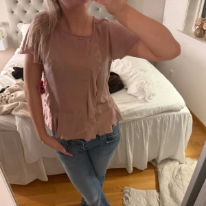 Beige blus med volangdetalj - Säljer en söt beige blus med korta ärmar och volangdetalj framtill. Blusen har en avslappnad passform och rund halsringning. Perfekt att styla med jeans eller kjol för en trendig look.