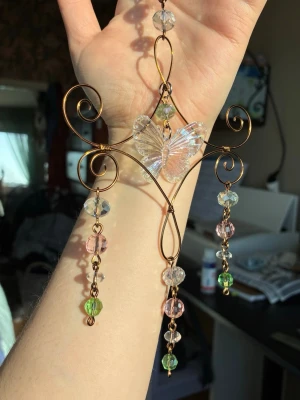 Solfångare med fjäril - Unikt hängsmycke i metalltråd med en stor, transparent fjäril i mitten och färgglada glaspärlor i grönt, rosa och klart. Smycket har snirkliga detaljer och tre hängande kedjor med pärlor. Perfekt som statement-accessoar eller dekoration. Fångar solen på ett vackert sätt. 
