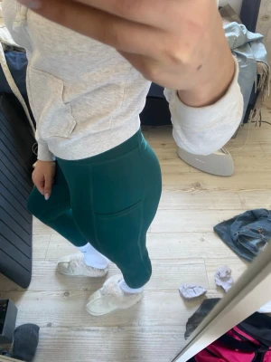 Mörkgröna leggings med fickor - Säljer ett par mörkgröna leggings med hög midja och praktiska sidofickor. De är tighta och stretchiga, perfekta för en avslappnad och sportig look. Materialet känns mjukt och följsamt, troligen i en blandning av polyester och elastan.