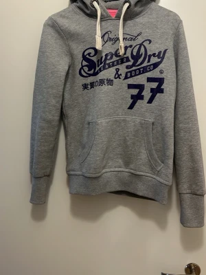 Grå hoodie från Superdry - Snygg grå hoodie från Superdry med mörkblå tryck på bröstet och japanska tecken. Hoodien har vit dragsko i huvan och en stor magficka. Perfekt för en avslappnad och trendig stil.