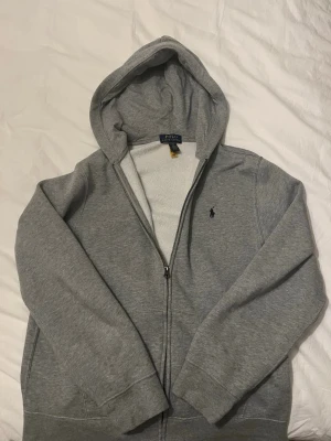 Grå hoodie från Polo Ralph Lauren - Snygg grå hoodie från Polo Ralph Lauren med dragkedja och klassisk huva. Tröjan har en liten broderad logga på bröstet och är tillverkad i mjukt bomullsmaterial. Perfekt för en avslappnad och stilren look. Nästan oanvänd, köpte den från kidsbrandstore och har endast användt den ett fåtal gånger. Passar perfekt till personer mellan 165 cm -175 cm