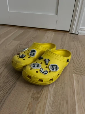 Gula Crocs med charms - Säljer ett par knallgula Crocs med flera olika charms på ovansidan, bland annat bokstäver och en giraff. Skorna är i klassisk Crocs-modell med rundad tå och rem bak. Perfekta för dig som vill sticka ut med färg och personliga detaljer.
