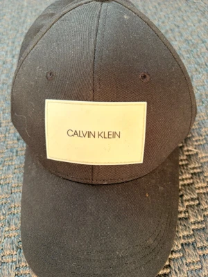 Svart keps från Calvin Klein - Snygg svart keps från Calvin Klein med justerbar snapback och vit logotyp framtill. Tillverkad i bomullstyg och har broderad text 'SWIMWEAR' på sidan. Perfekt accessoar för en stilren och avslappnad look.