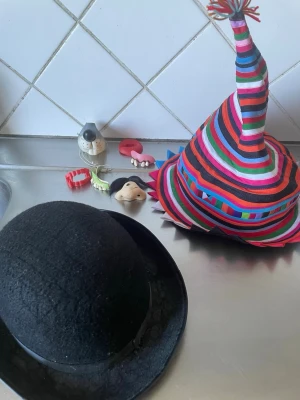 Maskeradkit med hattar och tillbehör - Säljer ett maskeradkit med en svart rundad hatt, en färgglad randig clownhatt med tofs, samt flera roliga tillbehör som lösnäsa, lösmun, löständer och lös mustasch. Perfekt för maskerad eller cosplay där du vill sticka ut och skapa en unik look.