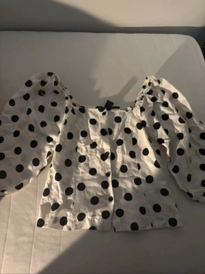 Vit prickig blus med puffärmar - Supersöt vit blus med stora svarta prickar och puffiga långärmar. Toppen har en rak, croppad passform och resår vid axlarna för en trendig look. Perfekt för dig som vill sticka ut med ett lekfullt mönster och voluminösa ärmar.