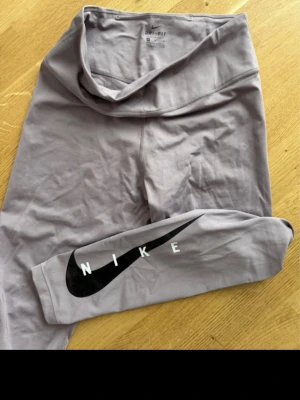 Ljuslila leggings från Nike - Snygga ljuslila leggings från Nike med hög midja och svart Nike-logga längs ena benet. Tillverkade i ett stretchigt och mjukt material som sitter skönt på kroppen. Perfekta för träning eller chill dagar.