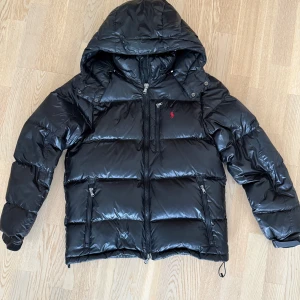 Svart pufferjacka från Polo Ralph Lauren - Svart glansig pufferjacka från Polo Ralph Lauren med huva, dragkedja framtill och två sidofickor med dragkedja. Jackan har röd broderad logga på bröstet och justerbar dragsko nertill. Perfekt nu när kylan smyger sig på. Size S, tts. Skick 9/10, medkommer tags samt påse. Pris 3999 nypris strax över 6k. 