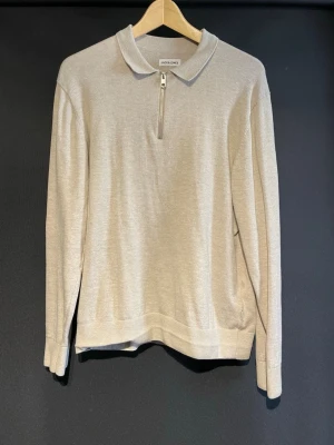 Beige stickad tröja med dragkedja - Stilren beige stickad tröja från Jack & Jones med krage och kort dragkedja framtill. Tröjan har långa ärmar, ribbade muddar och är tillverkad i ett mjukt, tunt stickat material. Perfekt för en avslappnad men snygg look. Pris kan diskuteras!