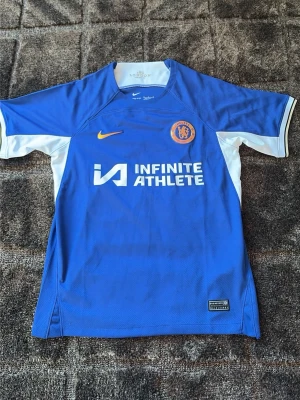 Chelsea matchtröja Palmer 20 Nike - Chelsea FC matchtröja i blått och vitt med Palmer 20 på ryggen. Tillverkad i lätt och ventilerande material, officiell logga och sponsortryck på bröstet. Perfekt för fotbollsträning eller att bära på matchdagar.