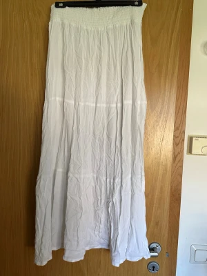 Vit långkjol med resår i midjan - Luftig vit långkjol med smockresår i midjan. Kjolen har en enkel och flowy design som passar perfekt till varma dagar. Tillverkad i ett tunt och mjukt material som ger en avslappnad känsla.
