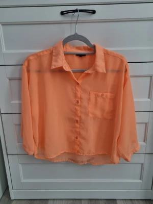 Persikofärgad transparent blus - Säljer en persikofärgad, transparent blus med krage och knappstängning framtill. Blusen har en bröstficka och är lite kortare i modellen med trekvartsärm. Perfekt för en trendig och avslappnad look.