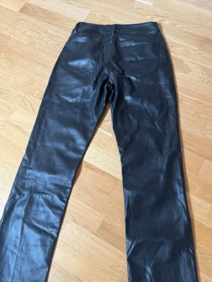 Svarta skinnbyxor med raka ben - Säljer ett par svarta skinnbyxor med raka ben och klassiska fickor både fram och bak. Byxorna har en snygg, glansig finish och är perfekta för dig som vill ha en cool och edgy look. Passar till många olika stilar och är ett riktigt statement-plagg.