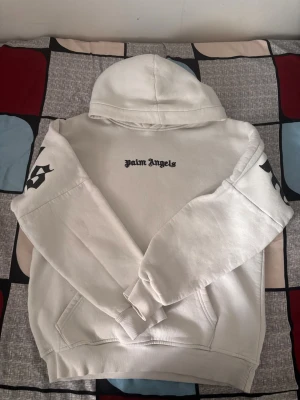 Vit hoodie från Palm Angels - Säljer en vit hoodie från Palm Angels med svart logotyp på bröstet och stora tryck på ryggen och ärmarna. Hoodien har ribbade muddar, känguruficka och en rejäl huva. Materialet är mjukt och bekvämt, perfekt för en avslappnad streetwear-look.