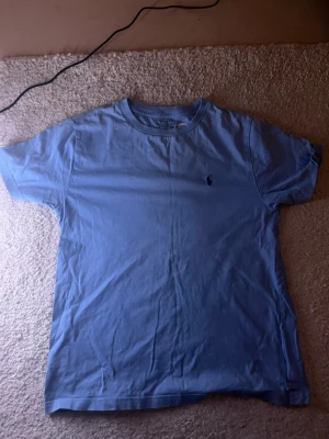 Blå t-shirt från Polo Ralph Lauren - Säljer en klassisk blå t-shirt från Polo Ralph Lauren med den ikoniska lilla loggan broderad på bröstet. T-shirten har rund halsringning och korta ärmar. Perfekt basplagg i mjuk bomull som funkar till allt.