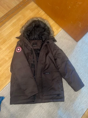 Brun dunjacka från Canadian Mountain - Mörkbrun dunjacka från Canadian Mountain med avtagbar huva och fuskpäls. Jackan har ett rött logomärke på ärmen, dragkedja och knappar framtill samt flera fickor. Fodrad insida för extra värme, perfekt för kalla vinterdagar. Storlek 160.