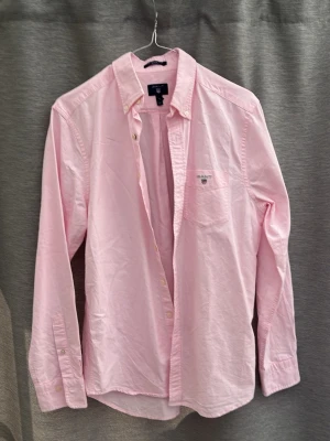 Ljusrosa skjorta från GANT - Säljer en ljusrosa skjorta från GANT med klassisk krage och knappar hela vägen fram. Skjortan har en bröstficka med broderad GANT-logga och långa ärmar med manschettknappar. Perfekt för dig som gillar stilrena plagg.