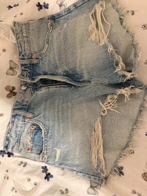 Ljusblå slitna jeansshorts - Trendiga ljusblå jeansshorts med medel midja och råa, fransiga kanter. Shortsen har slitningar framtill och klassiska fickor både fram och bak. Perfekta för sommaren och ger en avslappnad streetstyle-look. Köpte på vinted men passade inte. Säljs även på andra appar så skynda och fynda!