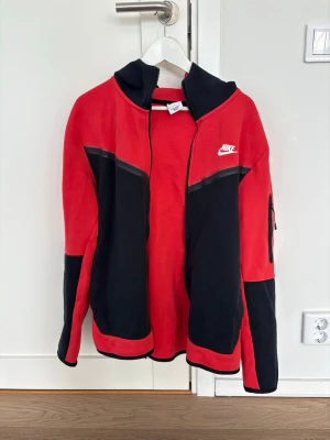Nike Tech Fleece kofta - Snygg röd och svart Tech Fleece från Nike med dragkedja framtill och vit logga på bröstet. Hoodien har svarta detaljer på ärmar och huva samt en ficka med dragkedja på ena ärmen. Perfekt för en sportig och avslappnad stil. Nypris ca 1500kr. Pris kan diskuteras vid snabb affär.