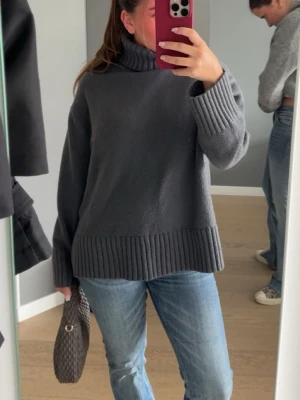 Mörkgrå stickad polotröja - Mysig mörkgrå stickad polotröja med breda ribbade muddar vid ärmslut och nederkant. Tröjan har en hög polokrage och en avslappnad passform, perfekt för lager-på-lager. Passar till jeans eller kjol för en chill look.