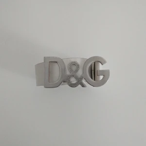 Vitt bälte med D&G-spänne - Snyggt vitt bälte med ett stort D&G-spänne i silver. Perfekt för att ge en stilren touch till din outfit. Bältet är tillverkat i ett elegant material och har en klassisk design. Varan är 110 cm lång😊 Kom med bud👍🏼