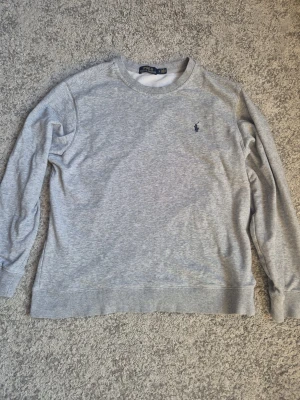 Grå sweatshirt från Polo Ralph Lauren - Klassisk grå sweatshirt från Polo Ralph Lauren med rund halsringning och broderad logga på bröstet. Tröjan har långa ärmar, ribbade muddar och är gjord i mjukt bomullsmaterial. Perfekt för en avslappnad och stilren look. Storlek L men passar bäst på M.