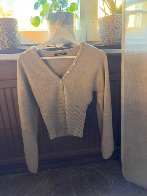 Beige kofta från Gina Tricot - Säljer en beige kofta från Gina Tricot med V-ringning och dekorativa knappar framtill. Koftan har långa ärmar och ribbstickad nederkant som ger en snygg passform. Perfekt att slänga över en topp för en chill look. Nästan helt oanvänd!!