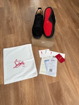 Svarta Louboutin sneakers med röd sula - Snygga svarta sneakers från Christian Louboutin med ikonisk röd sula. Skorna har en klassisk låg design, svarta skosnören och är tillverkade i mocka och läder. Perfekta för dig som vill sticka ut med exklusiv stil.Hör av dig vid minsta lilla