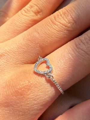 Silverring med hjärta och stenar - Snygg silverfärgad ring med ett öppet hjärta dekorerat med små gnistrande stenar. Ringen har en tunn, elegant band och hjärtat är i fokus. Perfekt accessoar för att lyfta din stil. Den kan justeras något baserat på storlek jag har omkrets 5,5cm på fingret. Testad.