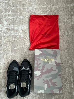 Valentino Garavani svarta sneakers - Snygga svarta sneakers från Valentino Garavani med grått och vitt kamouflagemönster. Skorna har ovandel i mesh och mocka, samt detaljer i läder. Klassisk låg modell med svarta snören och gummisula med nitar på hälen. Kommer med originalkartong och röd dustbag.
