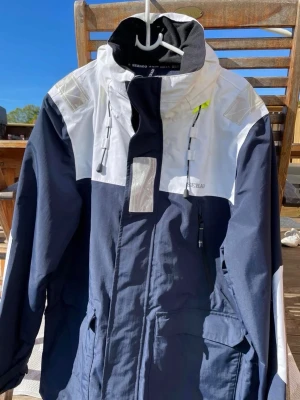 Blåvit vindjacka från Sebago - Snygg vindjacka från Sebago i marinblått och vitt med reflexdetaljer och hög krage. Jackan har dragsko i huvan, flera fickor och stängs med dragkedja. Perfekt för blåsiga dagar eller segling och har en sportig look.