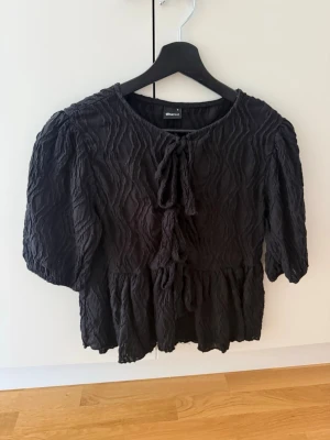 Svart blus med knyt från Gina Tricot - Snygg svart blus från Gina Tricot med strukturmönster och knytning fram. Blusen har volang nertill, puffiga trekvartsärmar och rund halsringning. Perfekt för dig som gillar trendiga och bekväma plagg.
