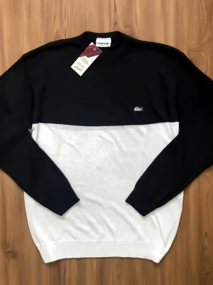 Svartvit stickad tröja från Lacoste - Snygg stickad tröja från Lacoste i svart och vitt med klassisk krokodillogga på bröstet. Tröjan har rund halsringning, långa ärmar och ribbade muddar. Perfekt för en stilren och modern look.