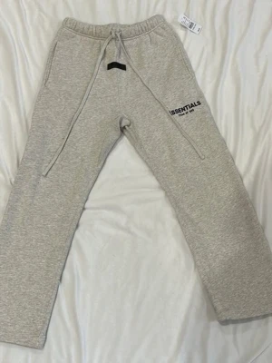 Grå mjukisbyxor från Essentials - Snygga grå mjukisbyxor från Essentials med svart logga på vänster lår. Byxorna har dragsko i midjan och två sidofickor. Perfekta för en chill och avslappnad stil. Tillverkade i mjukt material för extra komfort.