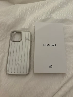Silverfärgat mobilskal från Rimowa 16 pro Max  - Snyggt och stilrent mobilskal från Rimowa i silverfärgad aluminium med klassiska räfflade linjer och logga längst ner. Skalet har en robust känsla och passar perfekt för dig som vill ha en exklusiv look på mobilen.