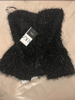 Svart paljettkorsett från Zara - Supersnygg svart korsettopp från Zara med paljetter och fluffig textur. Toppen är axelbandslös och har knappar framtill. Perfekt för dig som vill sticka ut och addera lite extra glam till din outfit.