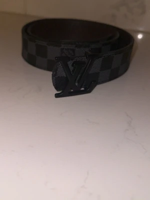 Svart rutigt bälte från M - Snyggt svart bälte från med klassiskt rutigt mönster och en cool -logga i metall som spänne. Perfekt för att lyfta din outfit med en exklusiv touch. Materialet är skinn och bältet har en mörk färgton.
