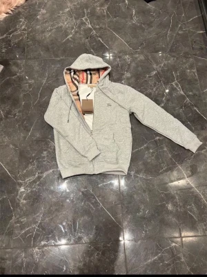 Grå zip-hoodie från Burberry - Säljer en grå zip-hoodie från Burberry med klassiskt rutigt foder i huvan och små diskreta logodetaljer på bröstet. Hoodien har dragsko i huvan, fickor framtill och ribbade muddar. Perfekt för en chill och stilren look.