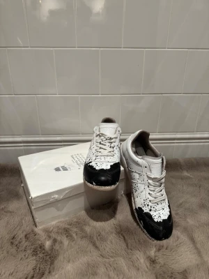 Maison Margiela vita och svarta sneakers - Unika vita sneakers från Maison Margiela med svart färgstänk och svart tåparti. Lågt skaft, klassisk siluett och vita snören. Tillverkade i läder med beige sula. Perfekta för dig som vill sticka ut med en kreativ och trendig look.