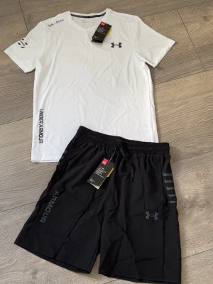 Under Armour träningsset svart/vit - Snyggt träningsset från Under Armour med en vit t-shirt och svarta shorts. T-shirten har tryckt logga på bröstet och text längs sidan. Shortsen har elastisk midja, dragsko och logga på benet. Perfekt för gym eller löpning.