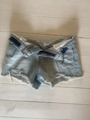 Ljusblå jeansshorts från Zara - Säljer ett par ljusblå jeansshorts från Zara med råa, fransiga kanter och slitna detaljer. Klassisk femficksmodell med dragkedja och knapp. Perfekta för varma dagar och en avslappnad look.