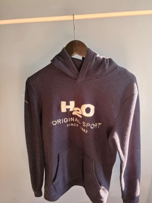 Mörkblå hoodie från H2O - Mysig mörkblå hoodie från H2O med broderad logga och texten 'Original Sport Since 1982' på bröstet. Tillverkad i ett mjukt, fleece-liknande material och har en klassisk känguruficka framtill samt huva.