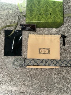 Gucci bälte med GG-mönster och silver-spänne - Säljer ett snyggt Gucci bälte med klassiskt GG-mönster i blått och beige. Bältet har ett stort, rundat silverfärgat spänne med Gucci-logga. Materialet är skinn och syntet, och bältet kommer med originalpåse, box och certifikat. Storlek: 115cm