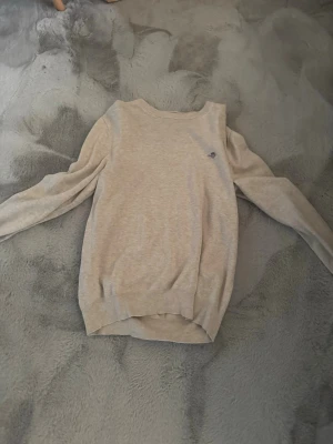 Beige stickad tröja från Gant - Snygg beige stickad tröja från Gant med rund halsringning och diskret logga broderad på bröstet. Tröjan har långa ärmar och ribbade muddar vid ärmslut och nederkant. Perfekt basplagg till garderoben.