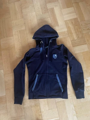 Svart hoodie med dragkedja från Kingsland - Snygg svart hoodie med dragkedja framtill, huva med breda snören och två fickor med dekorativa band. På bröstet finns ett litet emblem. Tillverkad i mjukt bomullsliknande material, perfekt för en avslappnad stil.