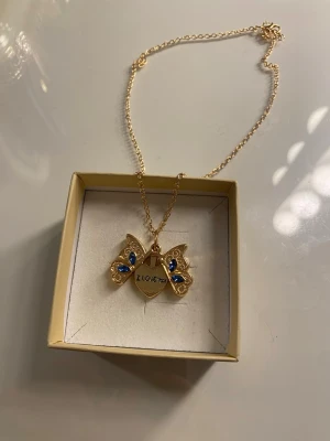 Guldigt halsband med blå stenar och fjäril  - Snyggt halsband i guldfärg med fjärilshänge dekorerat med blå stenar. Hänget kan öppnas och avslöjar ett hjärta med texten 'I LOVE YOU' inuti. Perfekt för dig som gillar detaljerade och unika smycken.