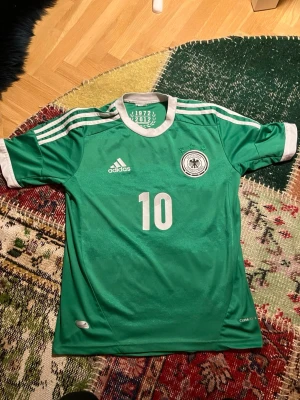 Grön Tyskland tröja Adidas Podolski - Säljer en grön matchtröja från Adidas med Tysklands landslagsemblem och nummer 10. Podolski står på ryggen. Tröjan har vita detaljer, tre ränder på axlarna och är tillverkad i ett lätt, ventilerande material. Perfekt för fotbollsträning eller att bära till vardags.